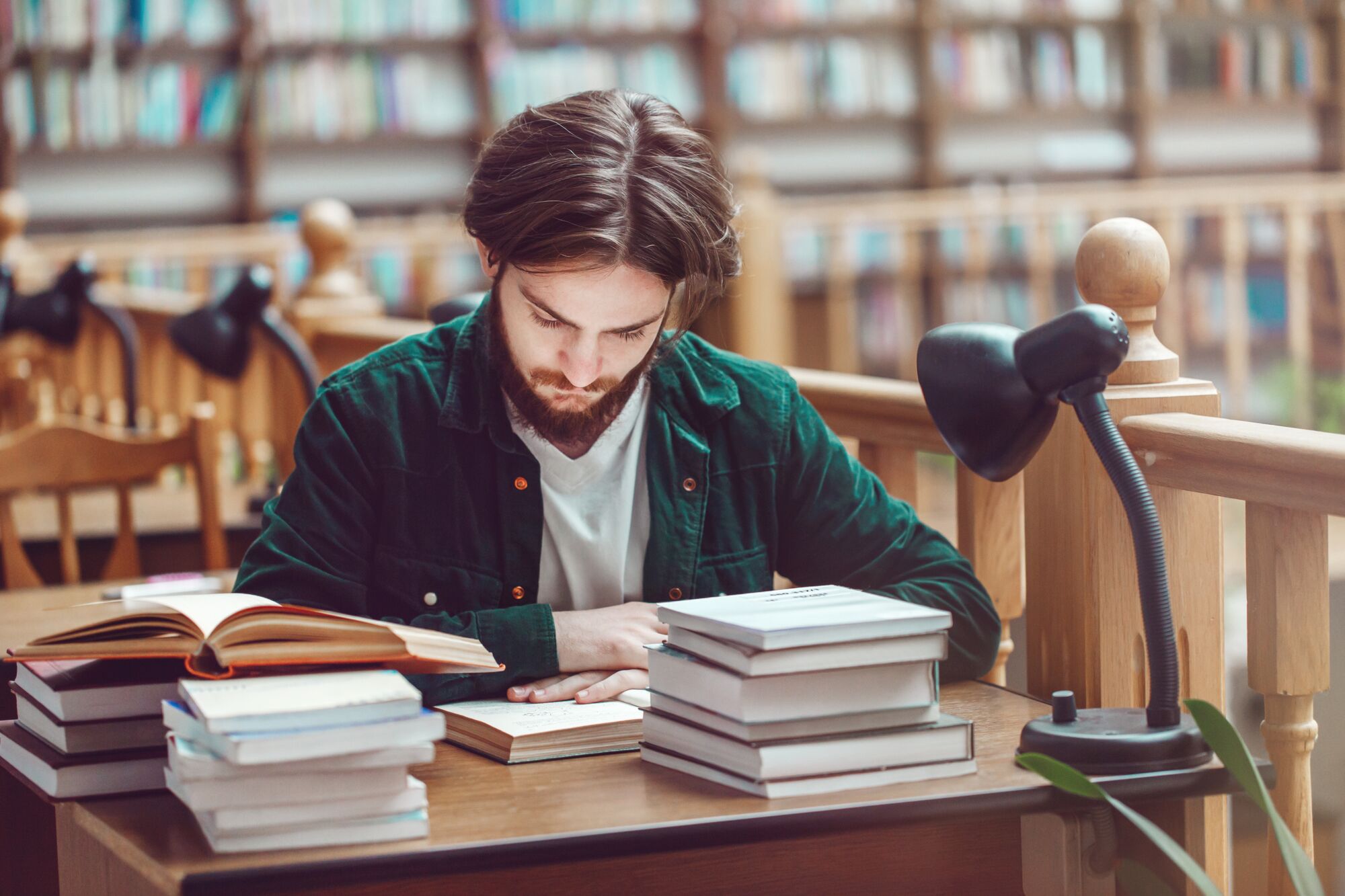 A young man studies intently at a library table, surrounded by stacked books and a desk lamp, emphasizing a focused learning environment.